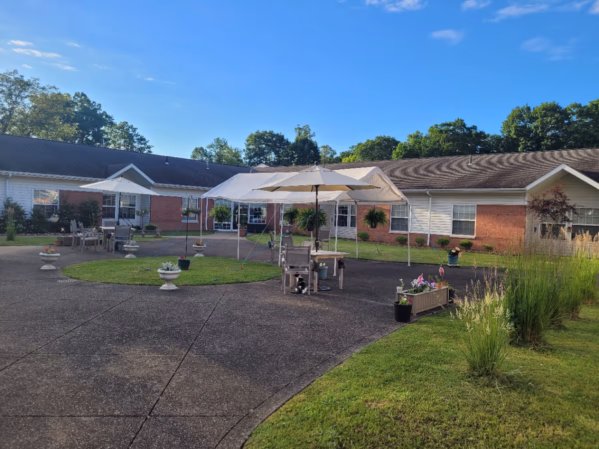 Outdoor courtyard area of a nursing and rehabilitation facility with paved walkways, green grass, potted plants, and patio tables with umbrellas. The building surrounding the courtyard has white siding and red brick accents under a clear blue sky.