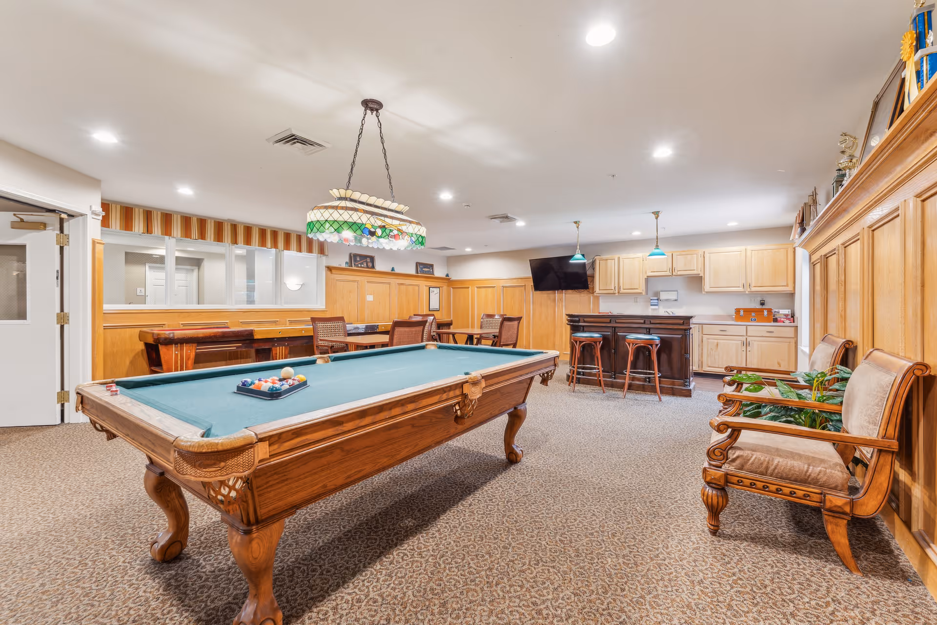 A spacious interior recreation room featuring a pool table under a stained-glass hanging light, a bar with stools, and seating along wood-paneled walls.