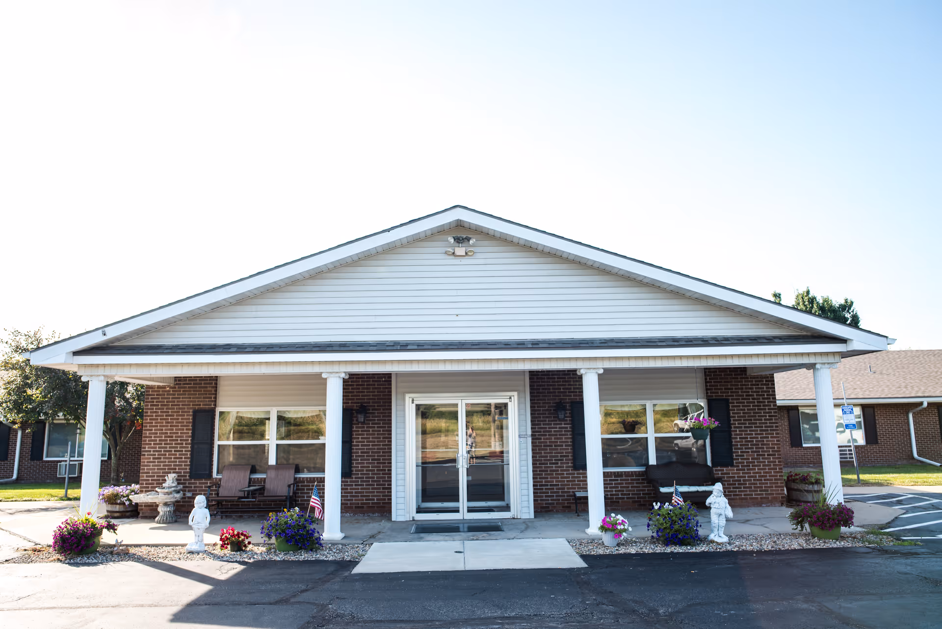 Front exterior view of a single-story brick building with white columns supporting a covered entrance. There are two windows on either side of the glass double doors, flower pots, small statues, and a bench on the porch area. The sky is clear and the surrounding area is well-maintained.