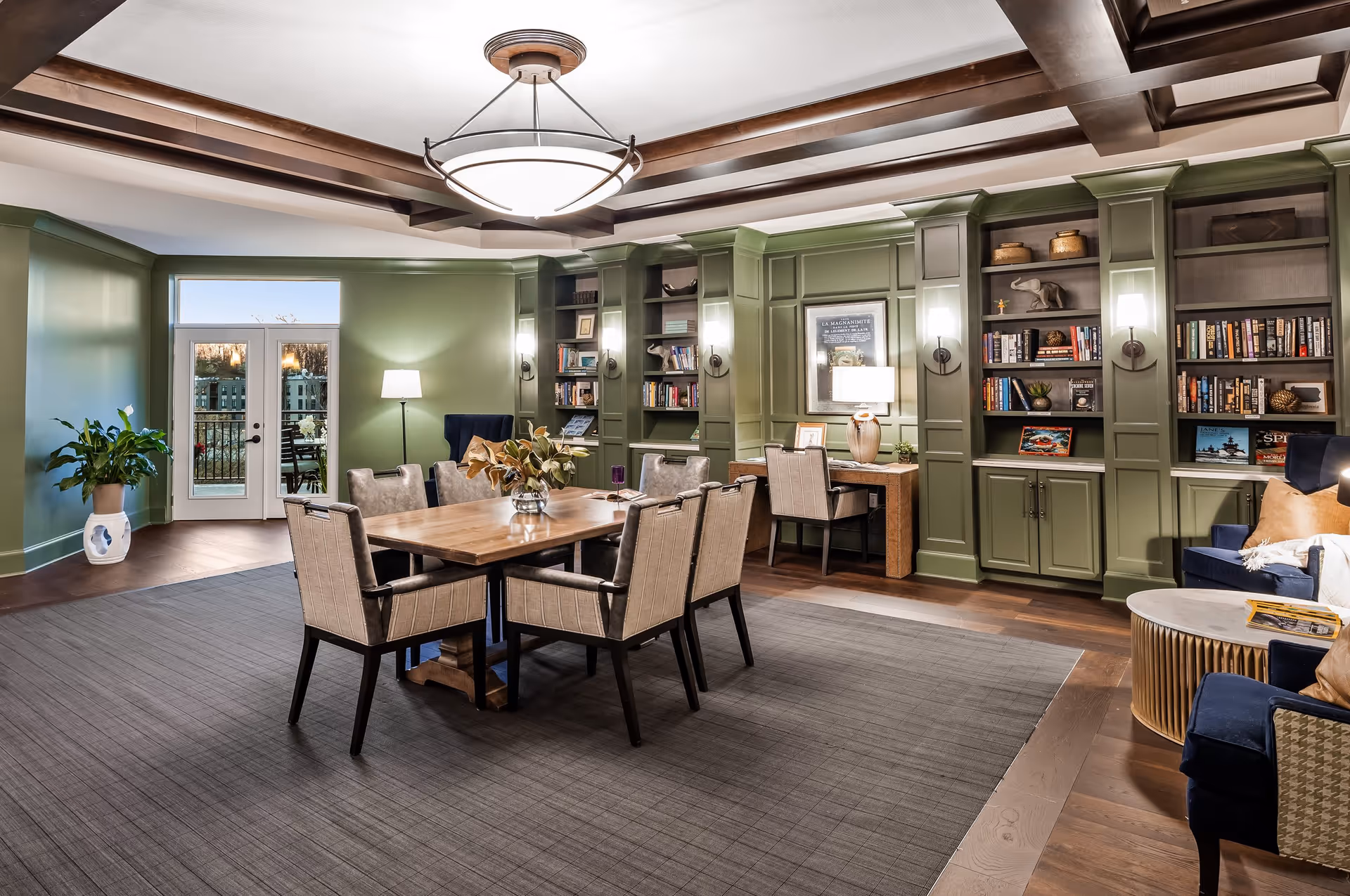 A well-lit interior room with green paneled walls featuring built-in bookshelves filled with books and decorative items. In the center, there is a wooden table surrounded by eight upholstered chairs. A large ceiling light fixture hangs above the table. To the right, there are two blue armchairs with tan pillows and a round coffee table. A desk with a chair and a lamp is positioned against the wall. Double glass doors lead outside to a patio area with outdoor seating. A potted plant is placed near the doors.