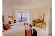 Bright bedroom with two twin beds featuring white and red bedding, a window with a view, a wooden vanity with a mirror, a potted plant, and light-colored walls.