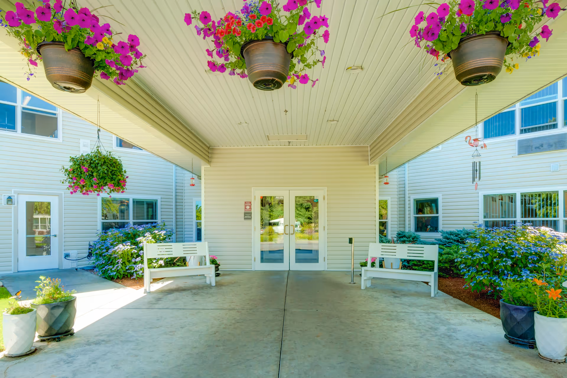 Covered entrance area of a building with two white benches on either side, hanging flower pots with purple and pink flowers, potted plants on the ground, and double glass doors in the center leading inside.