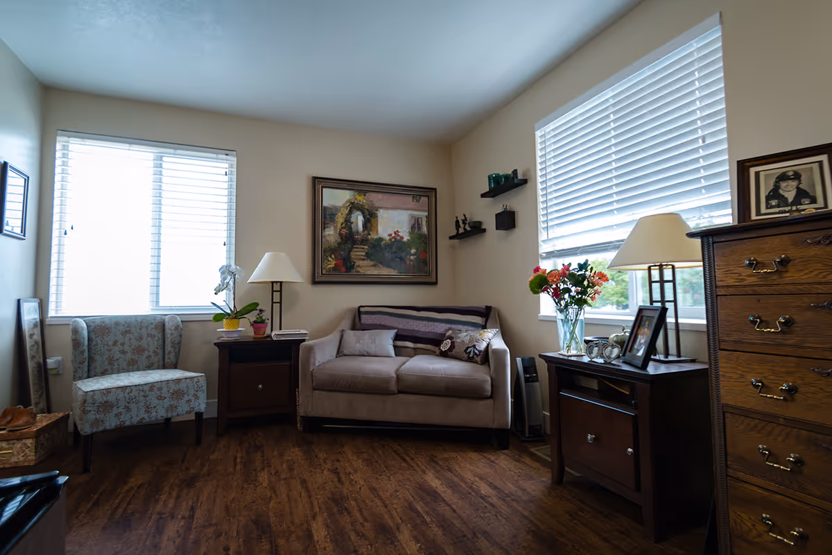A cozy living room with a beige loveseat adorned with pillows and a throw blanket, a floral patterned armchair, two wooden side tables with lamps, a vase of flowers, framed photos, and a wooden chest of drawers. Two windows with white blinds allow natural light into the room, and a painting hangs on the wall above the loveseat.