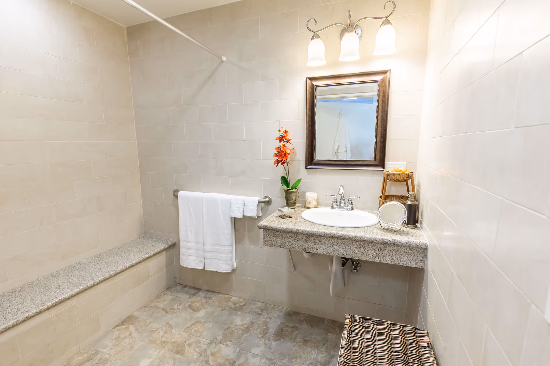 A clean bathroom with beige tiled walls and floor. There is a granite countertop with a sink, a mirror with a dark frame above it, and a light fixture with two bulbs. A white towel hangs on a metal bar on the wall, and a small orange flower arrangement is placed on the countertop. There is a built-in bench along one wall and a wicker basket on the floor.