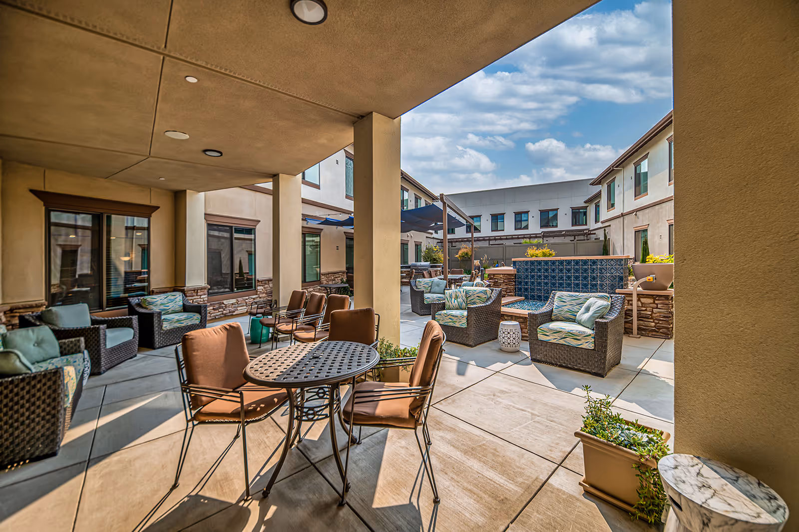 Outdoor patio area at Loma Clara Senior Living with multiple seating arrangements including cushioned wicker chairs and metal chairs around tables. The space is partially covered with a ceiling and has a blue tiled water feature. The area is surrounded by the building with windows and has potted plants and a clear blue sky with some clouds above.