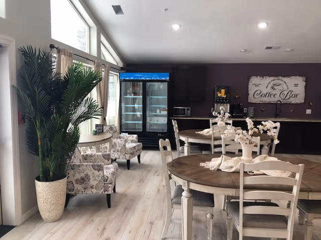 Interior view of a cozy common area in an assisted living center featuring round wooden tables with chairs, floral centerpieces, a coffee bar with a sign on the wall, a refrigerator, and two floral-patterned armchairs near large windows with curtains. A large potted plant is placed near the entrance.