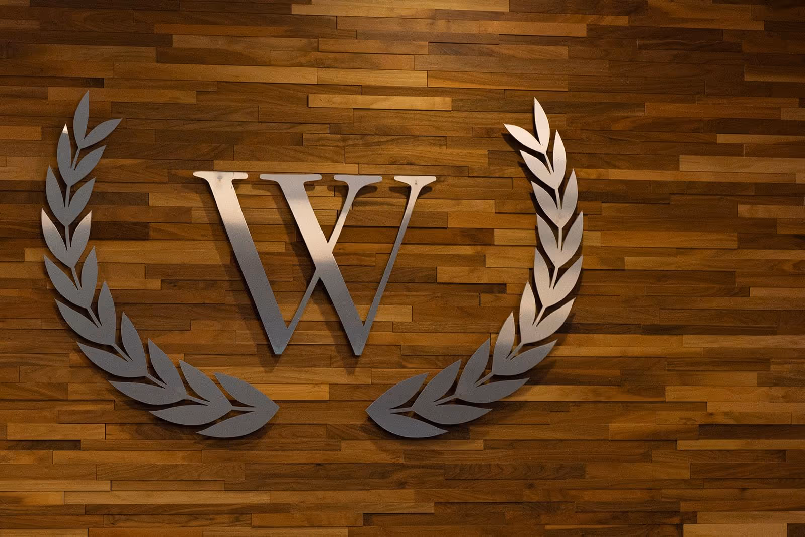 A metallic letter W mounted on a wooden wall, flanked by two stylized laurel branches on each side.