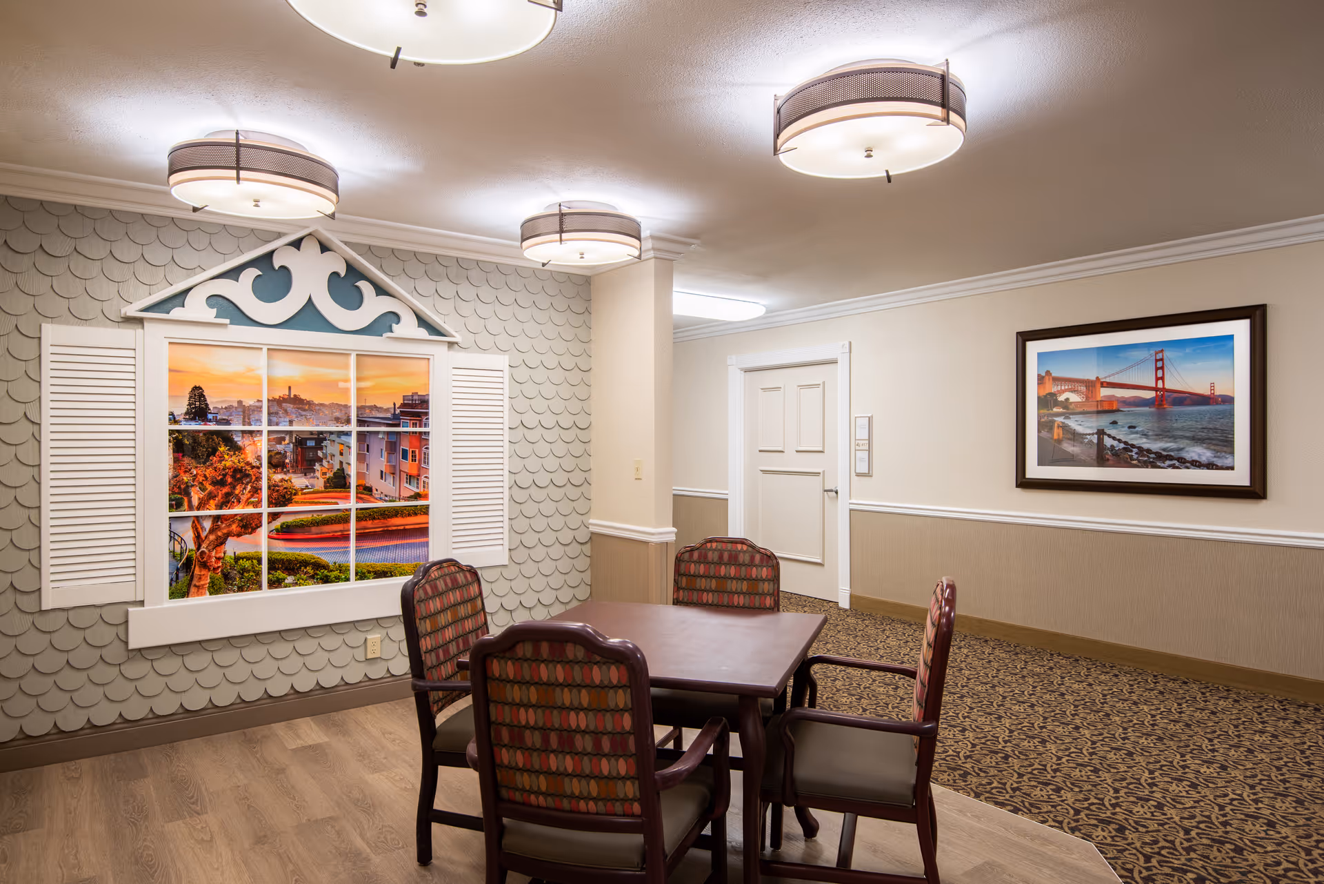 A cozy interior room with a square wooden table surrounded by four cushioned chairs with patterned upholstery. The walls are decorated with a faux window featuring a scenic view of a city at sunset and a framed picture of the Golden Gate Bridge. The ceiling has three round light fixtures, and the floor has a combination of wood and patterned carpet.