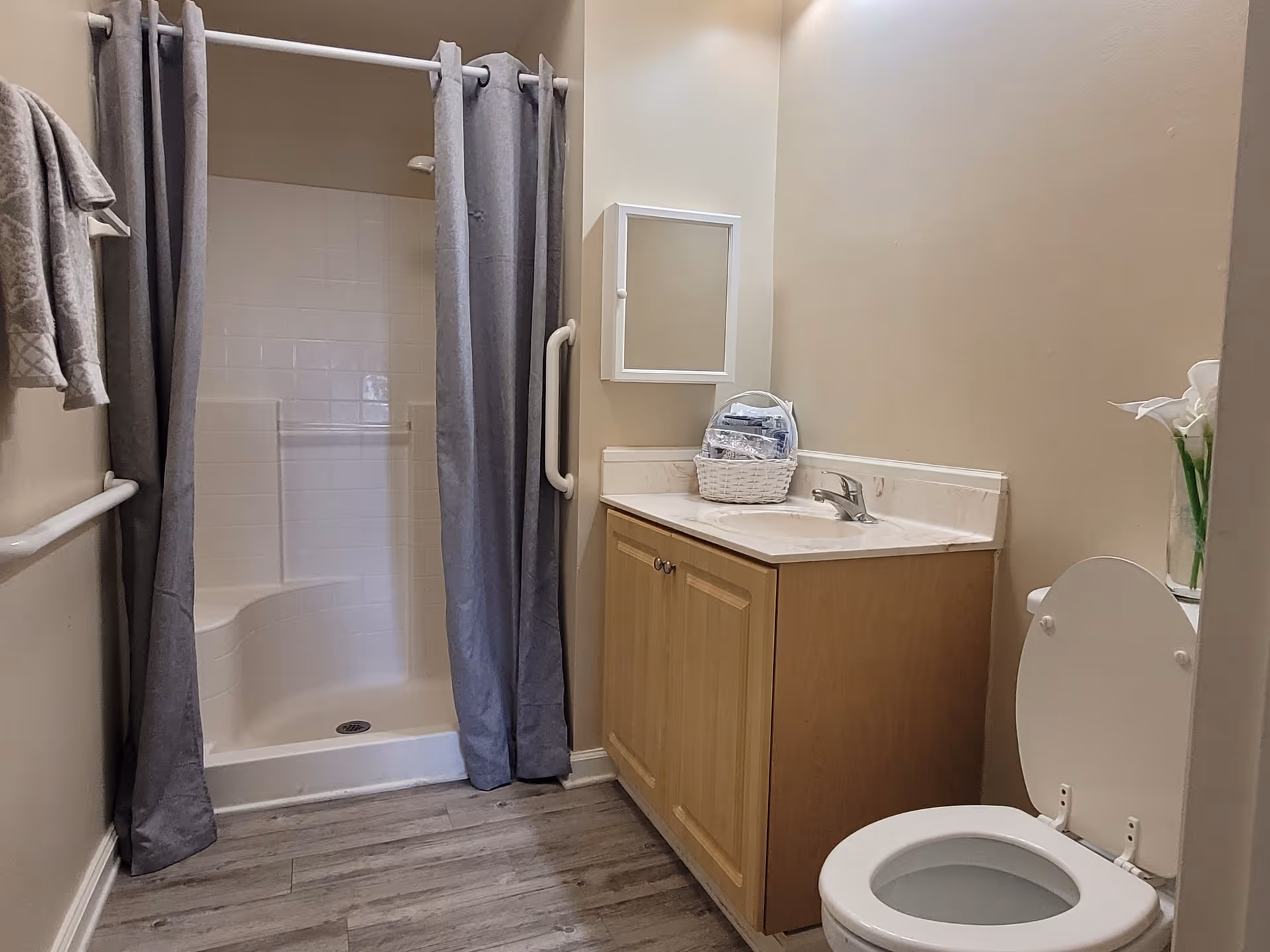 A clean bathroom featuring a walk-in shower with a gray curtain, a wooden vanity with a sink and a small basket of toiletries, a toilet with the lid open, and a vase with white flowers on the toilet tank. The walls are beige and the floor has wood-like tiles.