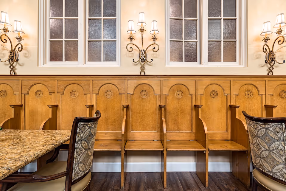 Interior view of a dining area with a wooden bench featuring individual seats and armrests along the wall, two upholstered chairs with patterned fabric, a granite table corner, three wall-mounted ornate light fixtures, and two windows with frosted glass panes.