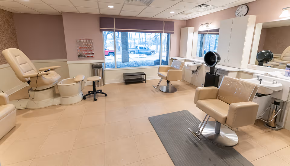 Interior view of a salon area in a senior living facility with beige salon chairs, a pedicure station, a nail polish display on the wall, large windows showing trees and parked vehicles outside, and mirrors with sinks and hair drying equipment.