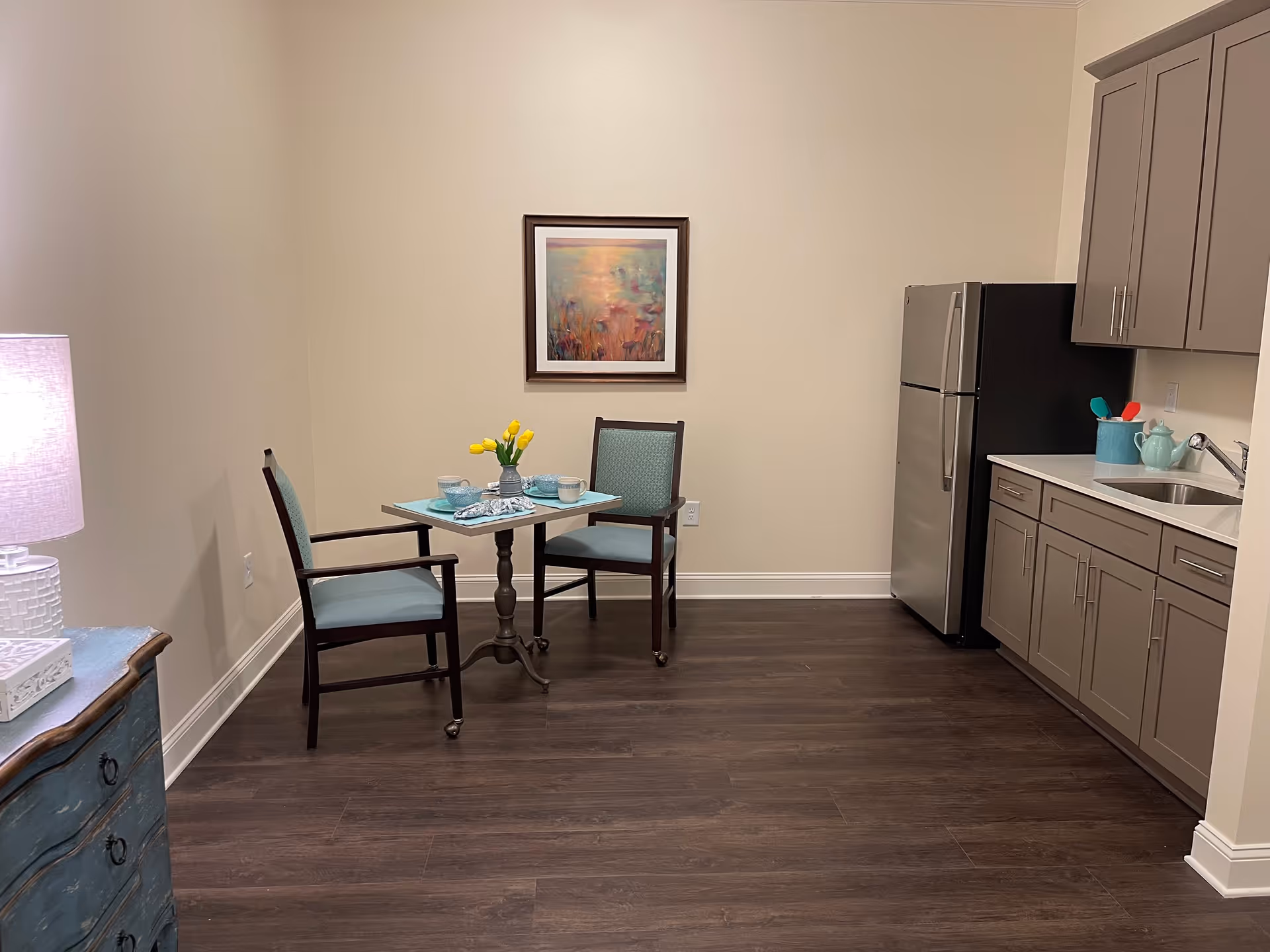 A small kitchen area with a stainless steel refrigerator, beige cabinets, and a sink. There is a small table set for two with blue placemats, bowls, mugs, and a vase of yellow tulips. A framed abstract painting hangs on the beige wall above the table. A blue chest of drawers with a white lamp is visible on the left side.
