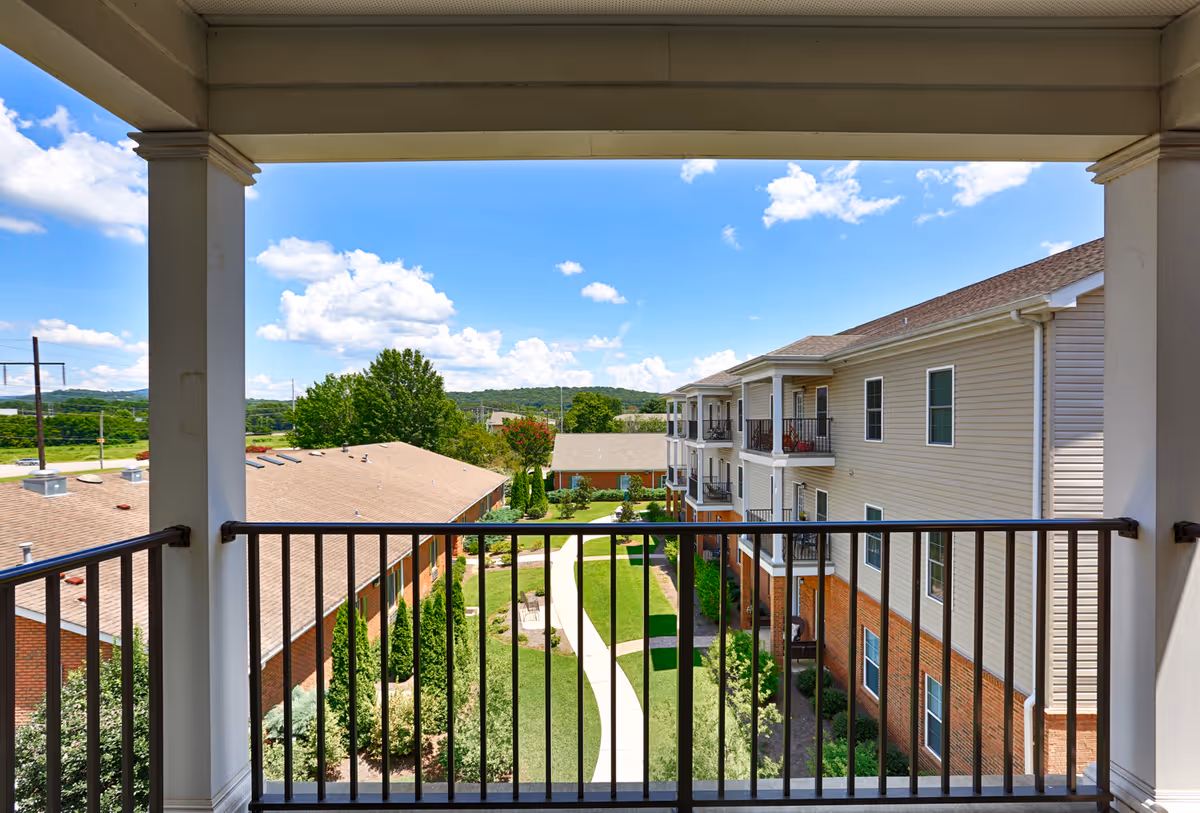 View from a covered balcony overlooking a landscaped courtyard with a walking path, surrounded by multi-story residential buildings under a blue sky with scattered clouds.