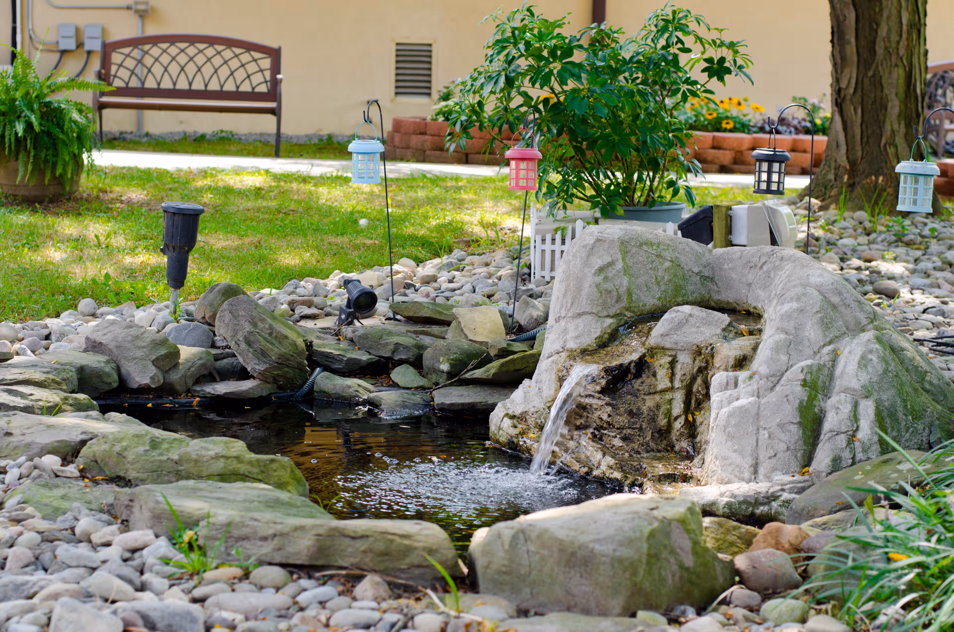 A small outdoor garden pond with a rock waterfall feature surrounded by stones and greenery. There are small decorative lanterns hanging on metal stakes around the pond, a bench in the background, and a building wall behind the garden area.
