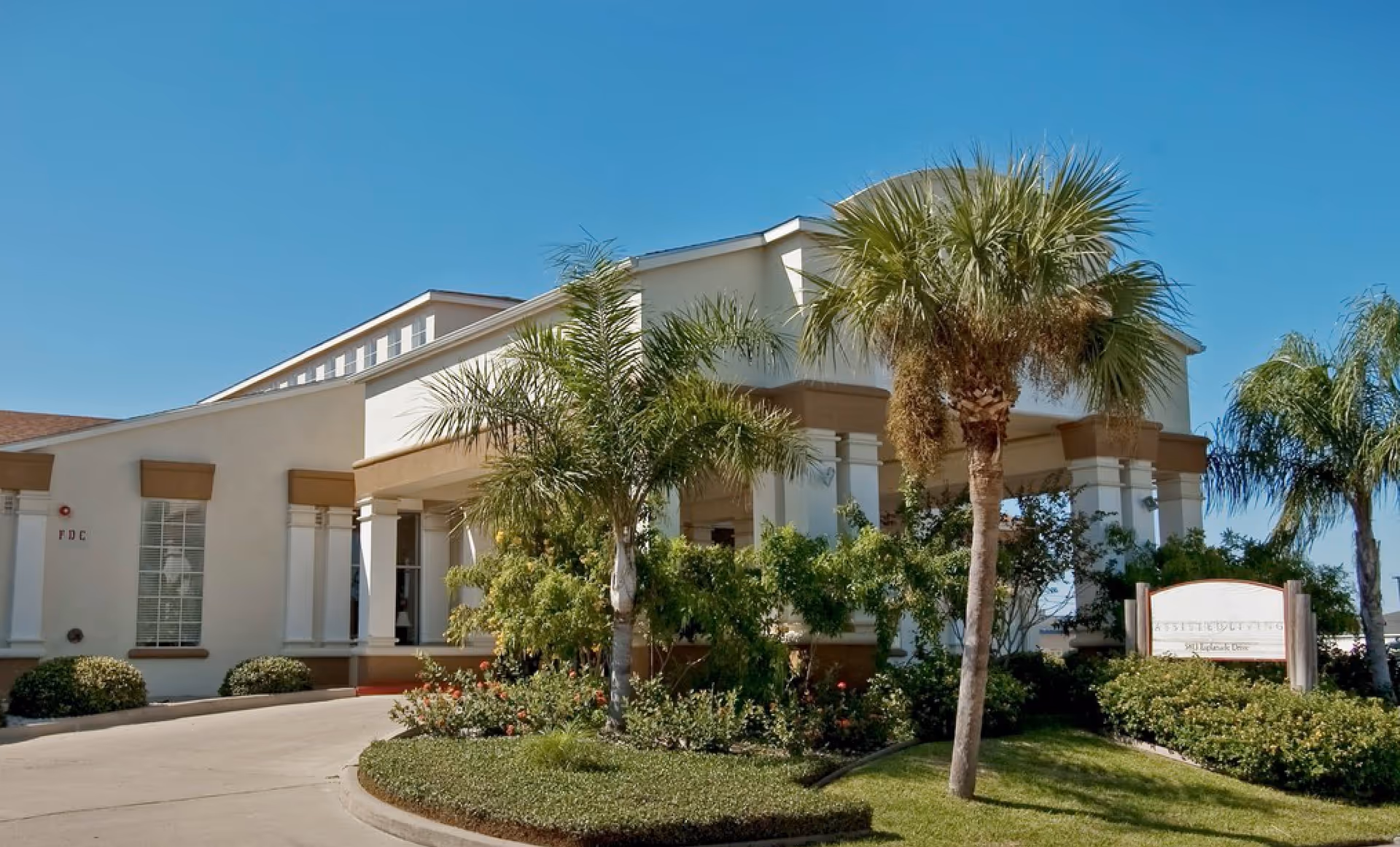Exterior view of The Waterford at Corpus Christi facility showing a beige building with white columns, surrounded by palm trees and well-maintained landscaping under a clear blue sky.