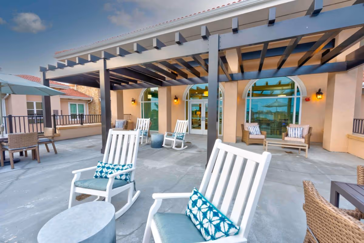 Outdoor patio area with white rocking chairs and wicker seating under a pergola in front of a building.