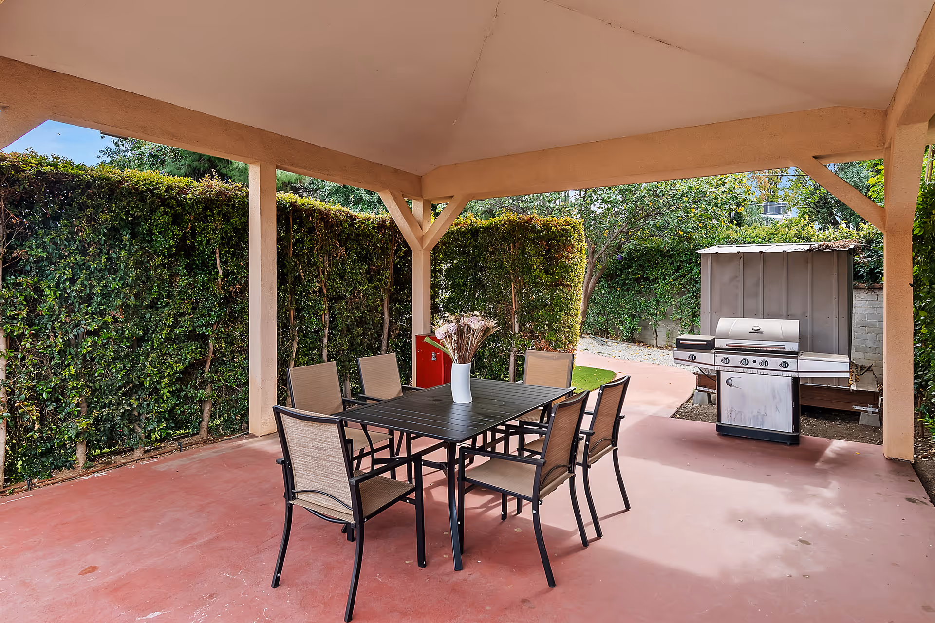 Covered outdoor patio with a dining table and six chairs and a gas grill next to hedges.