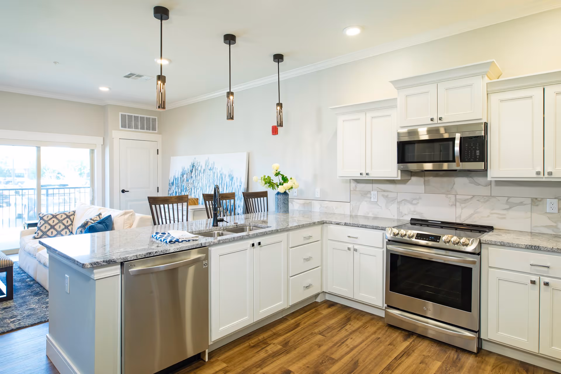 Bright and modern kitchen with white cabinets, stainless steel appliances including a dishwasher, oven, and microwave. Granite countertops with a double sink and three pendant lights hanging above. In the background, a dining area with wooden chairs and a sofa with decorative pillows near a sliding glass door leading to a balcony.