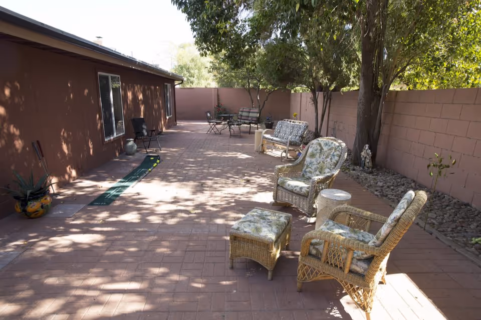 Outdoor patio area with brick flooring, featuring wicker chairs with floral cushions, a small matching ottoman, and a side table. There are additional seating arrangements with metal chairs and a bench along a brown stucco wall. Trees and plants provide shade and greenery along the perimeter.