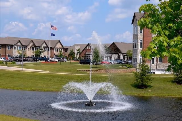 A fountain sprays water in a pond on landscaped grounds with senior living buildings and flags in the background.