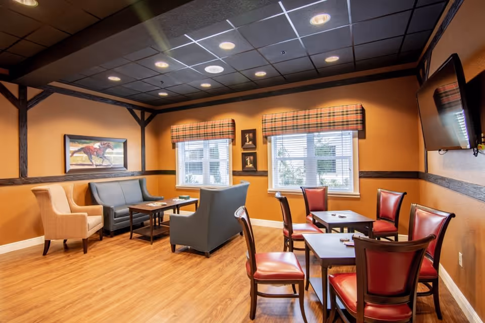A cozy common area with wooden flooring and warm orange walls featuring dark wood trim. The room has two windows with plaid valances, a wall-mounted TV, two small tables with red cushioned chairs, and a seating area with a gray sofa, loveseat, and a beige armchair. There is a painting of a horse on one wall and two small framed pictures between the windows.