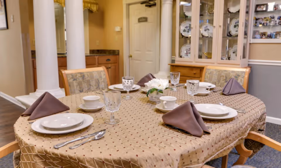 A dining table set for four with a beige tablecloth, brown folded napkins, white plates, cups, and crystal glasses. The background shows a china cabinet with decorative plates and a door, with two white columns and a countertop visible to the left.