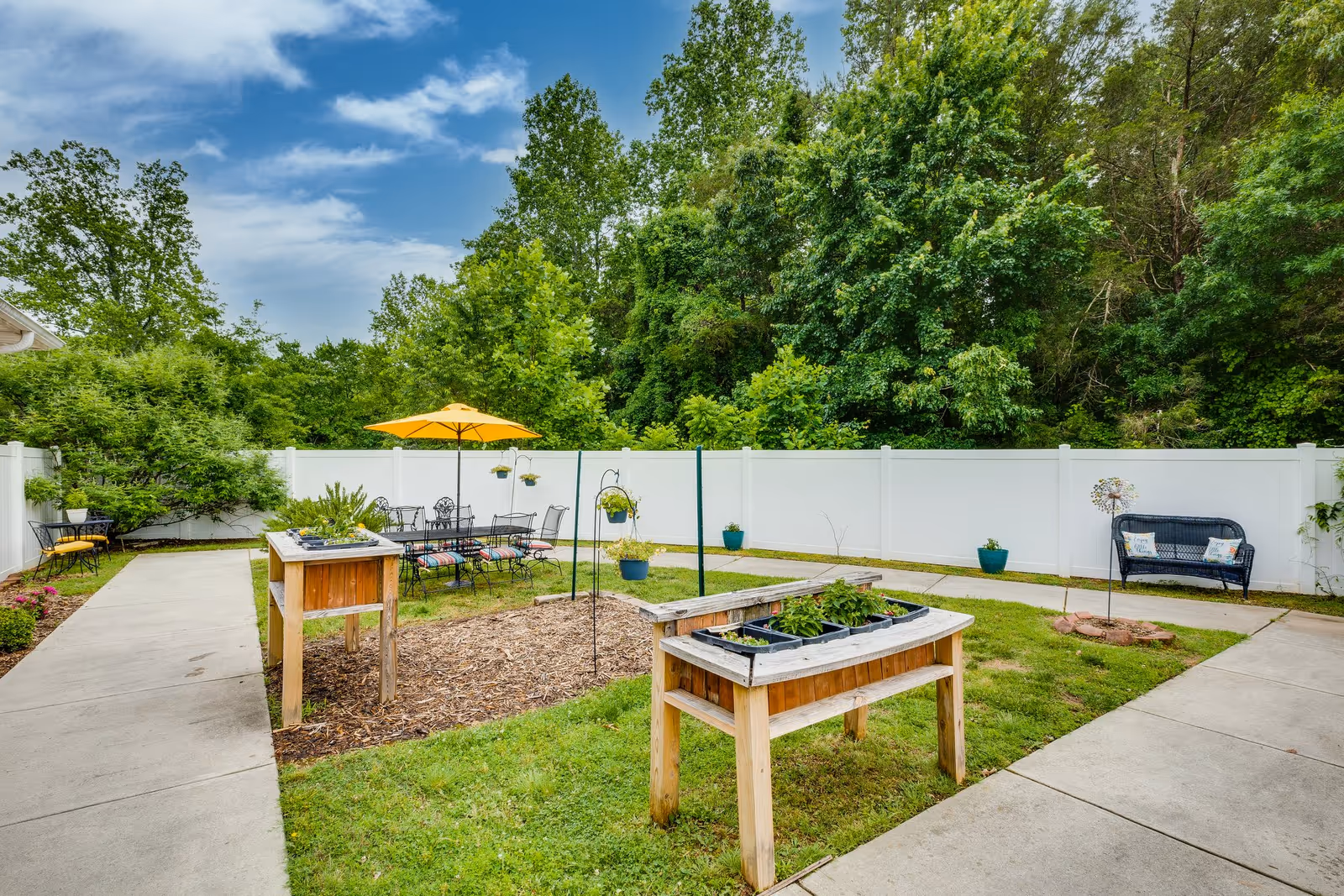 Outdoor garden area at Mallard Ridge Assisted Living with raised wooden planter boxes containing plants, a seating area with a table and chairs under a yellow umbrella, a black bench with decorative pillows, and a white privacy fence surrounding the space with trees and greenery in the background.