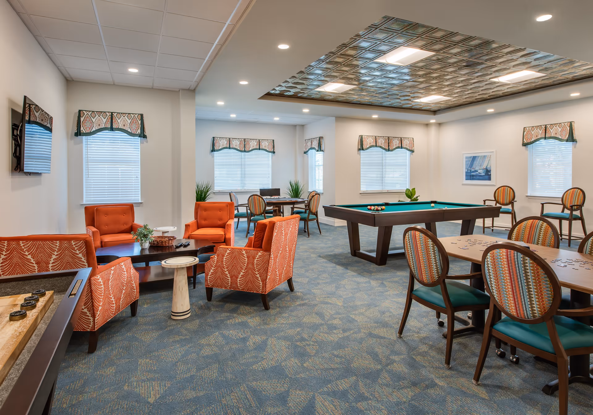 A spacious recreational room with a pool table, several tables with chairs, and a seating area with orange upholstered chairs and a small coffee table. The room has multiple windows with patterned valances, a decorative ceiling panel, and carpeted floors with a blue and beige pattern.