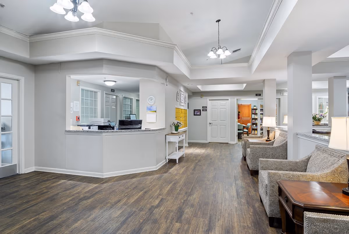 A bright and clean reception area in a senior living facility with a curved front desk, wood flooring, comfortable armchairs, side tables with lamps, and a hallway leading to other rooms. The walls are painted light gray, and there are ceiling lights providing illumination.