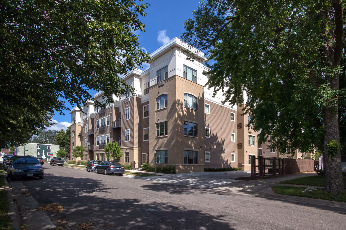 Exterior view of a multi-story residential building with a mix of brick and siding, surrounded by trees and parked cars along the street under a blue sky with some clouds.