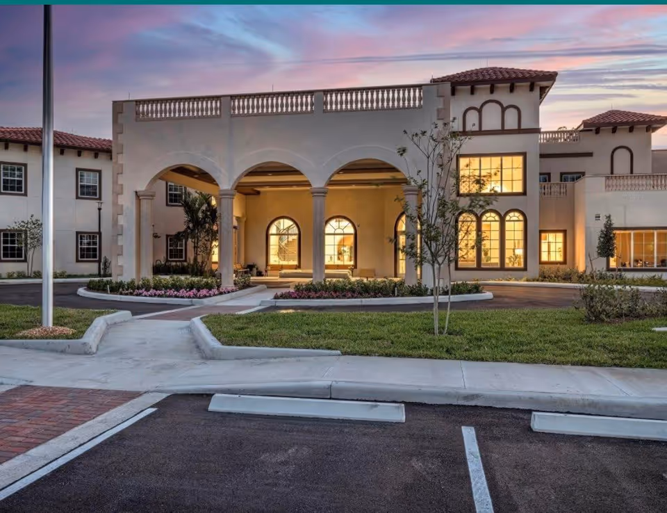 Front entrance of a Mediterranean-style building with an arched portico, lit windows, landscaping, and a parking area at dusk.