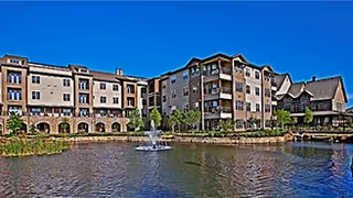 Exterior view of a multi-story senior living facility named Discovery Village At Southlake, featuring a large pond with a water fountain in the foreground and clear blue sky above.