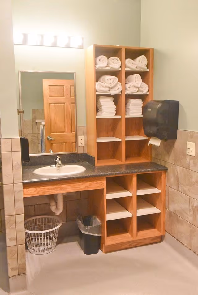 A bathroom vanity area with a sink, a large mirror, and wooden shelving filled with neatly folded white towels. There is a black paper towel dispenser mounted on the wall and a trash can and laundry basket underneath the countertop. The walls are tiled halfway up and painted light green above.