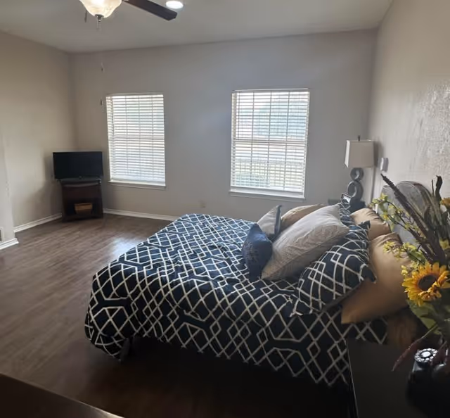 Sunlit bedroom with a bed dressed in a navy-and-white geometric comforter, two windows with blinds, a small TV on a stand, and a ceiling fan.
