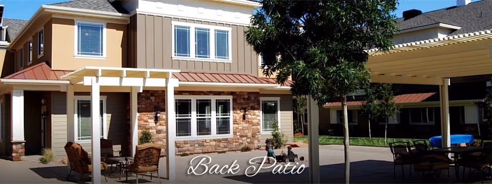 Outdoor patio area at a senior living facility with seating arrangements under pergolas, surrounded by a two-story building with windows and stone accents. A tree provides some shade in the patio area.