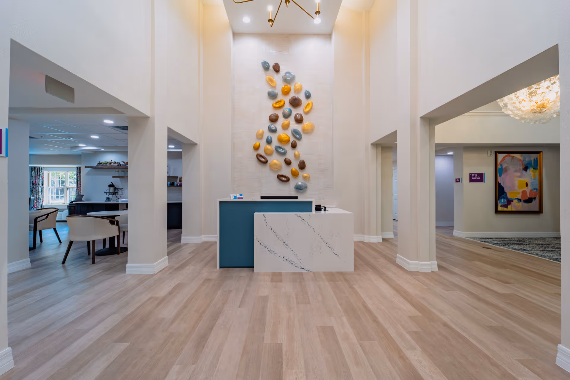 A spacious and modern reception area with light wood flooring and high ceilings. A white and teal reception desk is centered against a wall decorated with colorful, abstract wall art. To the left, there is a seating area with chairs and tables near a window with floral curtains. To the right, a hallway leads to other parts of the facility, with a colorful abstract painting on the wall.