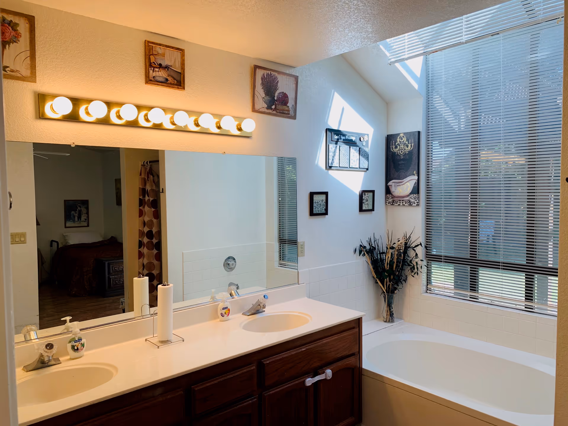 A bathroom with a double sink vanity featuring two faucets, a large mirror with bright vanity lights above, and a white countertop. To the right is a white bathtub next to a large window with blinds partially closed. The walls have decorative framed pictures, and a vase with dried plants is placed near the bathtub. A bedroom with a bed and nightstand is visible through an open doorway reflected in the mirror.
