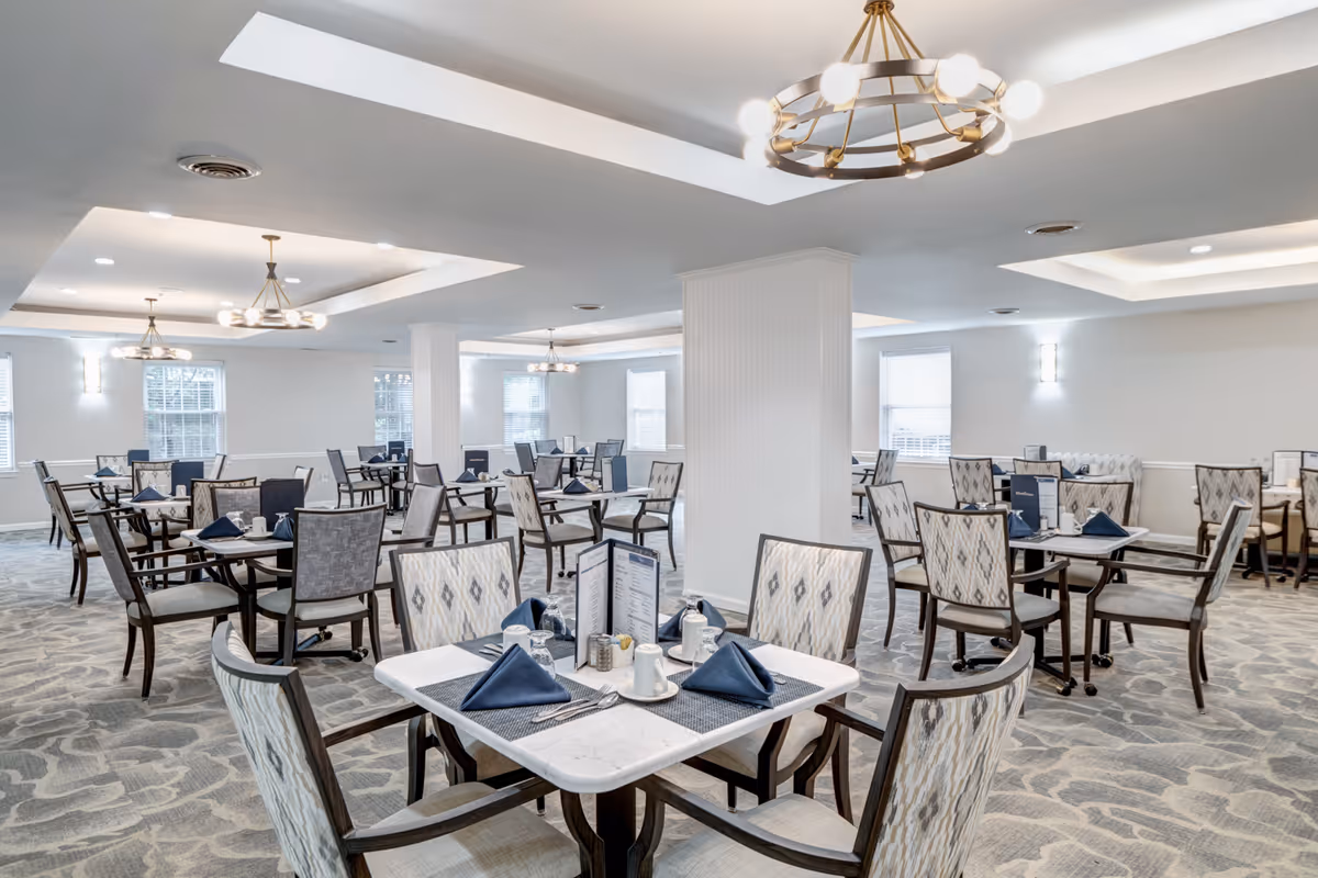 A spacious dining room with multiple tables set with blue napkins, menus, and white dishware. The room features patterned carpet, light-colored walls, large windows, and modern chandeliers hanging from the ceiling.