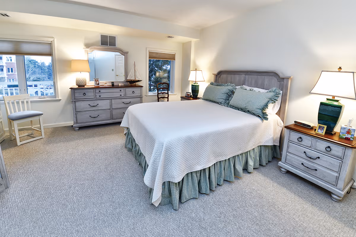 A bright and spacious bedroom with a large bed covered in a white quilt and green checkered pillows and bed skirt. The room features two windows with views of trees and buildings outside, a wooden dresser with a mirror, a chair, two bedside tables with lamps, and a carpeted floor.
