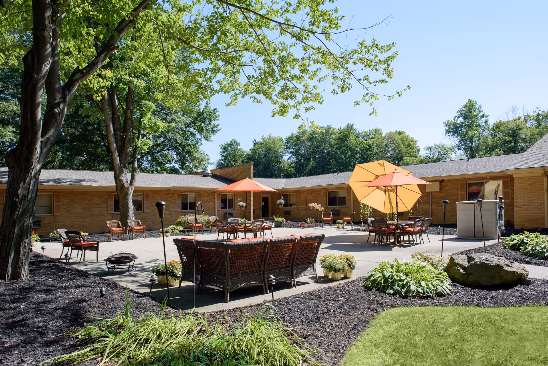 Sunny courtyard with patio seating, tables and umbrellas in front of a single-story brick building.