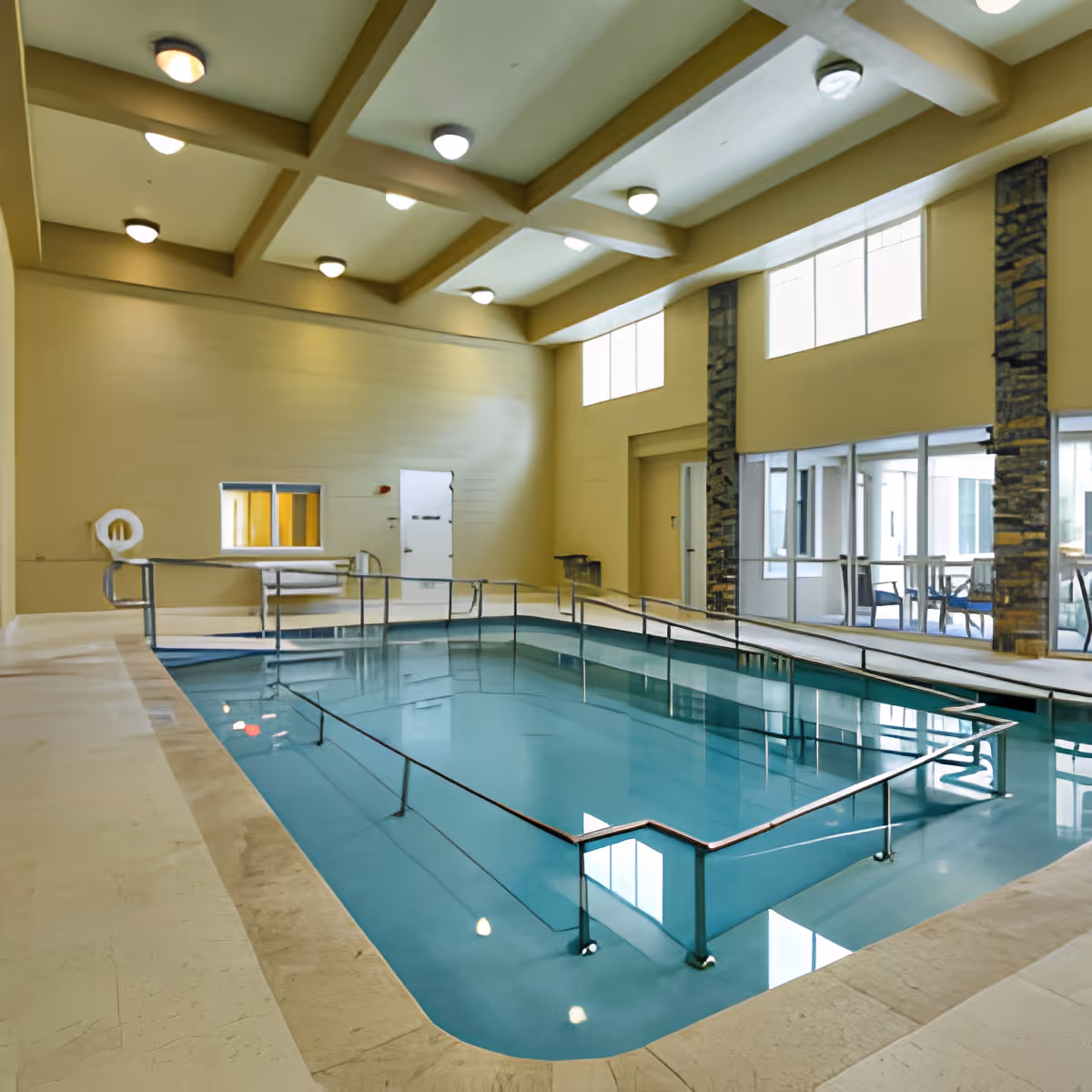 Indoor swimming pool with handrails for accessibility, surrounded by beige walls and large windows letting in natural light. The ceiling has multiple recessed lights and exposed beams. There is a seating area visible through glass doors on the right side.