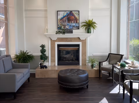 A cozy living room area with a fireplace centered on a beige tiled hearth. Above the fireplace is a framed picture of a cabin in the woods. On the hearth are decorative plants and a small dog figurine. To the left is a gray sofa, and to the right are two armchairs with a small round table between them. The room has large windows letting in natural light and dark wood flooring.