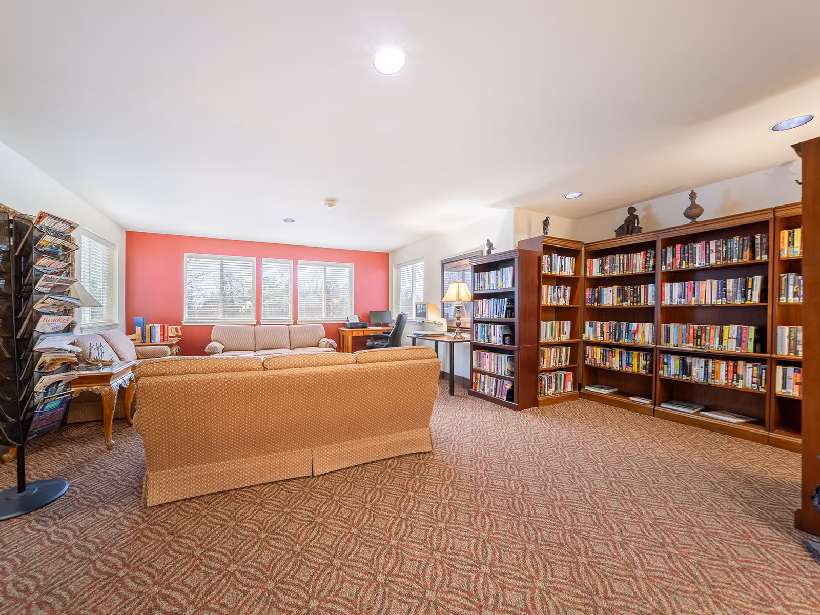 A cozy library or reading room with multiple bookshelves filled with books, two beige sofas facing each other, a small wooden side table with a lamp, a magazine rack, and two desks with computers near windows that let in natural light.