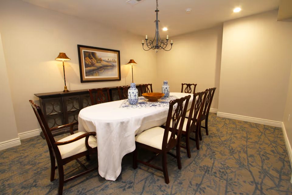 A dining room with an oval table covered by a white tablecloth and a decorative runner. The table is surrounded by eight wooden chairs with cushioned seats. On the table are two blue and white vases and a wooden bowl. Behind the table is a dark wooden sideboard with two lamps and a framed landscape painting on the wall. The room has beige walls, a patterned carpet, and a chandelier hanging from the ceiling.