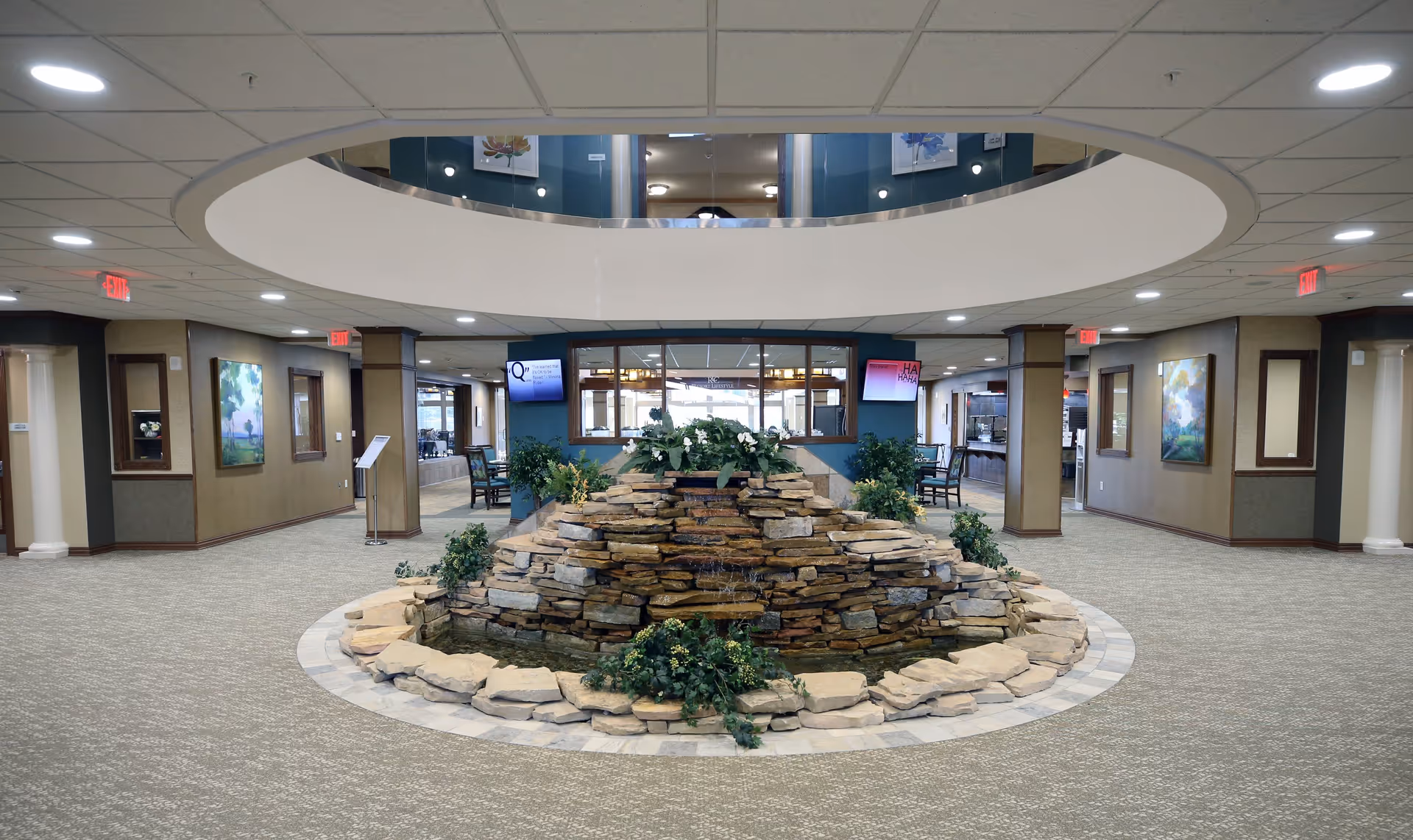 Interior view of a spacious common area in a senior living facility featuring a circular stone water fountain with greenery in the center. The area is carpeted and surrounded by walls with paintings and multiple doorways leading to other rooms. There are two digital display screens mounted on the walls behind the fountain, and the ceiling has recessed lighting with a large circular architectural feature above the fountain.
