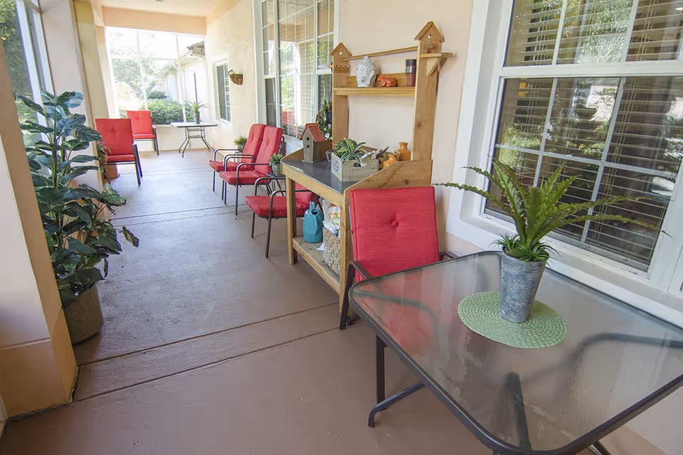 Covered outdoor patio area with several red cushioned chairs, a glass-top table with a potted plant, a wooden shelf with decorative items, and large windows with blinds.