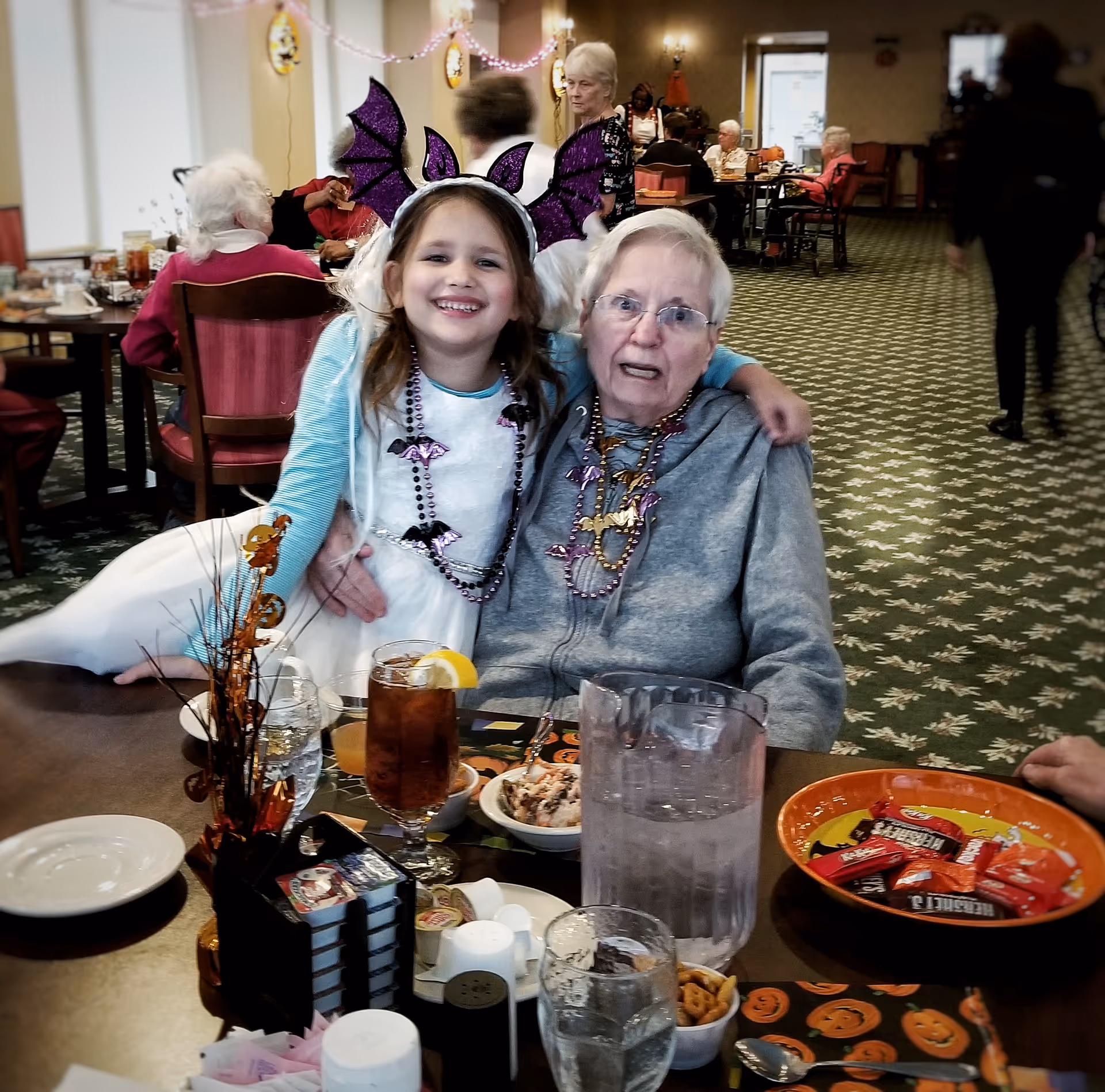 A young girl wearing a white dress and purple bat wing headband hugs an elderly woman seated at a table in a decorated dining room. The table has drinks, snacks, and Halloween-themed decorations. Other elderly people are seated and standing in the background.