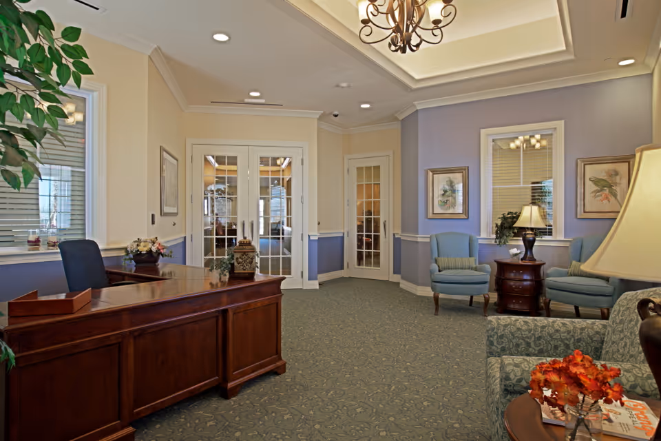 A well-lit reception area with a wooden desk and a blue office chair on the left. The room features a patterned carpet, light purple and beige walls, and two blue armchairs with a small round wooden table between them on the right. The table holds a lamp and a small plant. There are framed bird pictures on the walls and a chandelier hanging from the ceiling. Double glass doors and a single glass door are visible in the background.
