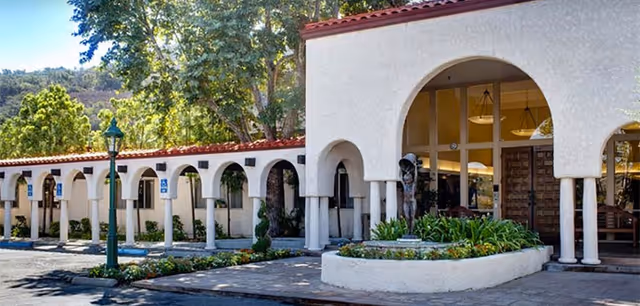 Arched stucco entrance and covered colonnade of a memory care building with planter beds and a tiled roof.