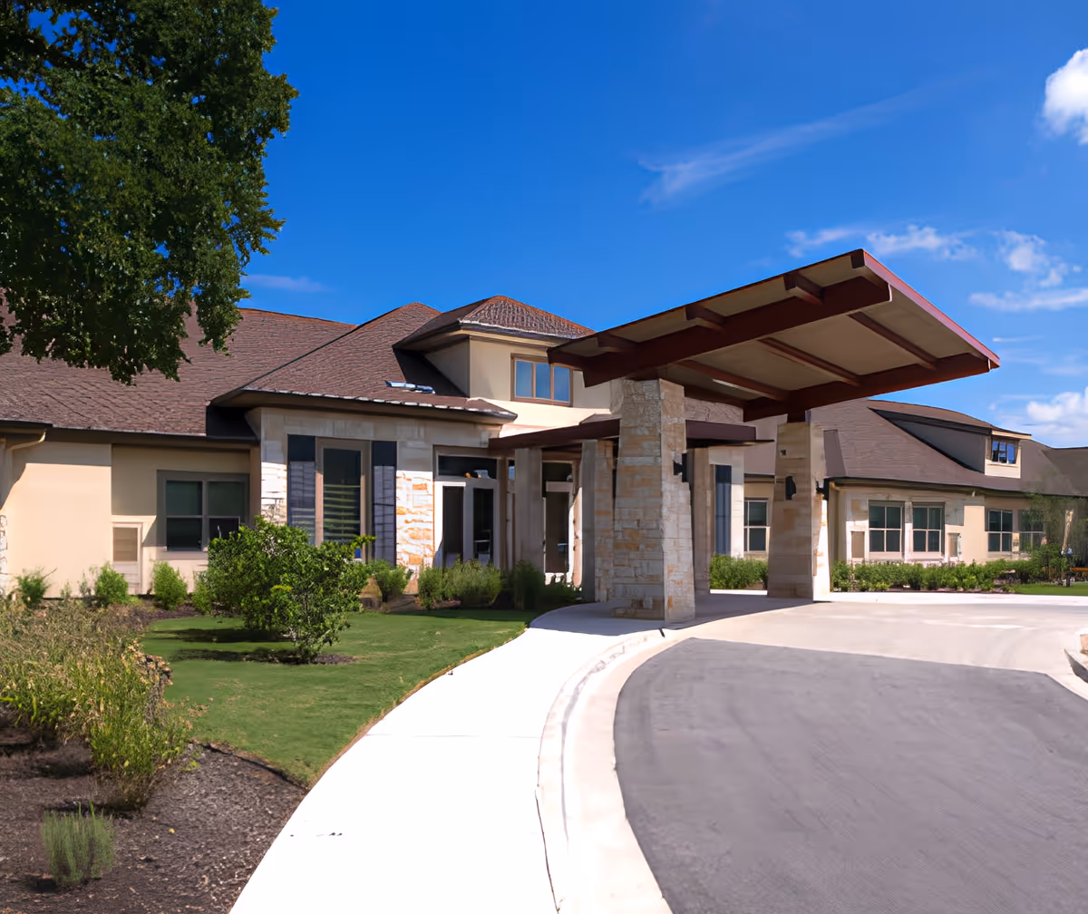 Exterior view of The Monarch at Round Rock facility showing a covered entrance with stone pillars, beige walls, multiple windows, a curved driveway, and landscaped greenery under a clear blue sky.
